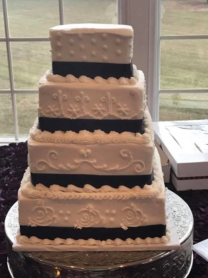 A multi-layer four-tier wedding cake with white icing and black ribbon accents, decorated with piped icing designs, placed on a decorative silver stand near a window.