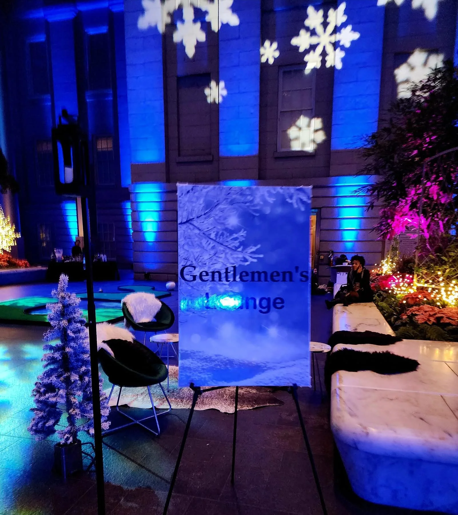 Decorative indoor space with holiday lights, snowflake projections, a sign reading "Gentlemen's Lounge," stylish chairs with fur accents, and bright decorative plants.