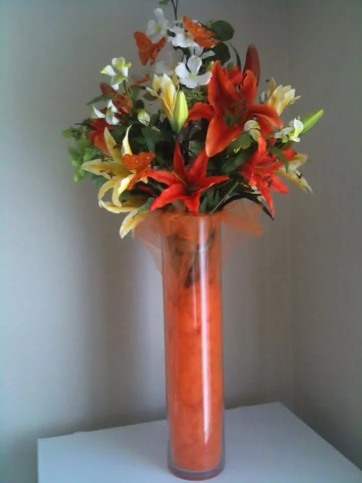 A tall glass vase filled with an orange-colored substance, holding a vibrant bouquet of white, yellow, orange, and red lilies and some greenery.