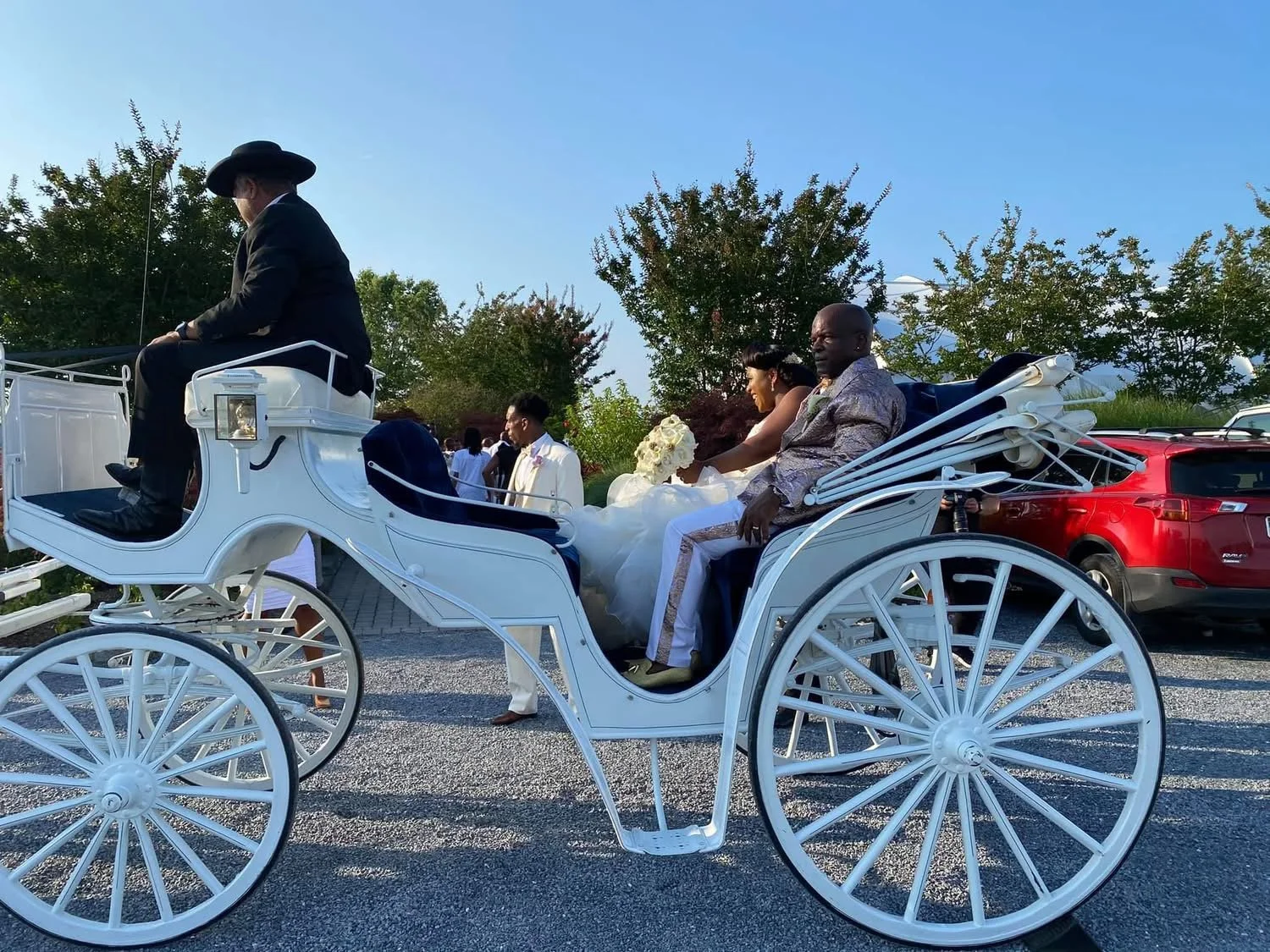 Couple dressed in wedding attire riding in a white horse-drawn carriage at an outdoor wedding event.