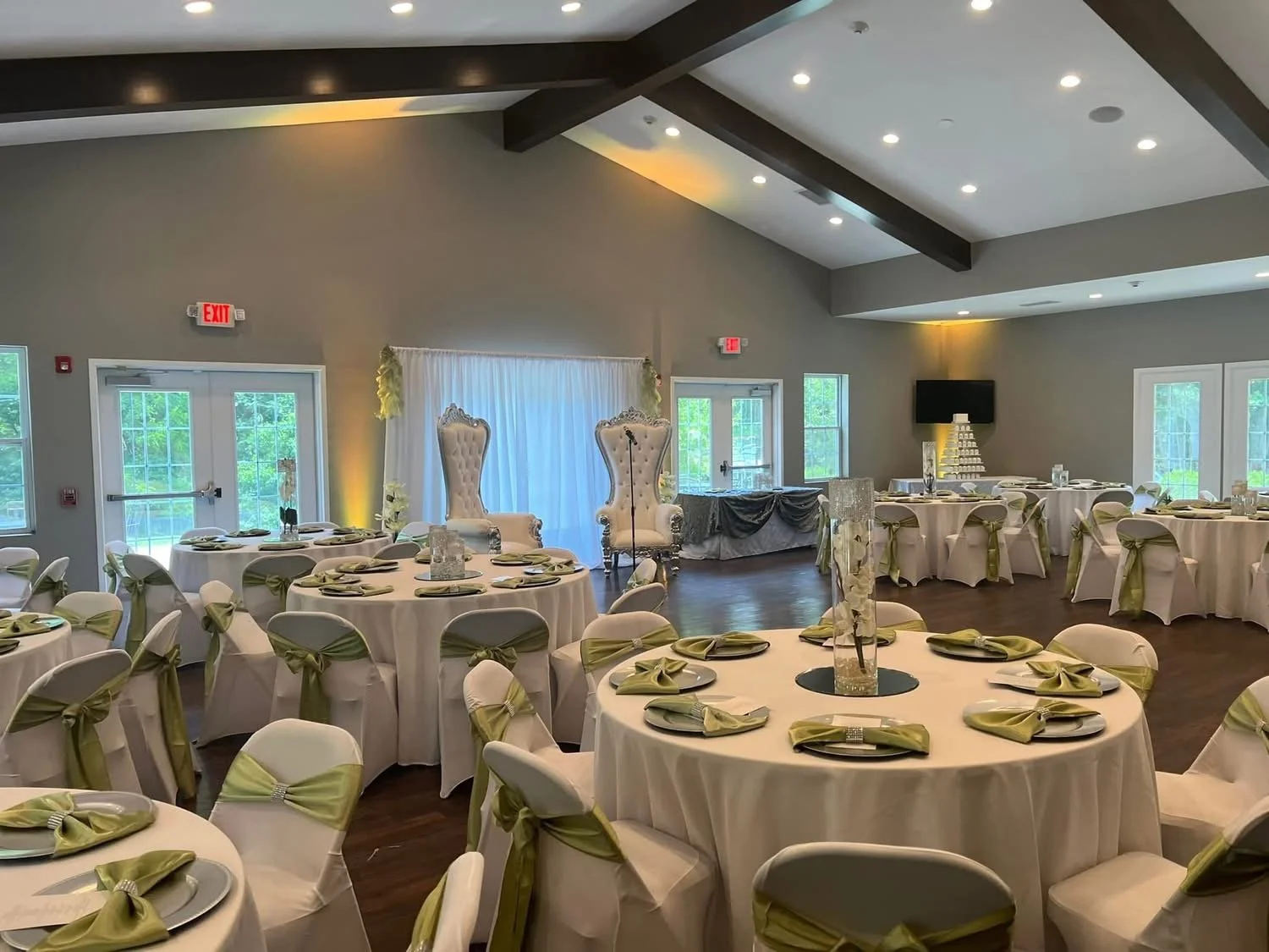 A banquet hall decorated for a wedding reception with round tables covered in white tablecloths, each with green napkins and plates, and white chairs adorned with green sashes. At the front, there are two tall, ornate high-back chairs and a backdrop 