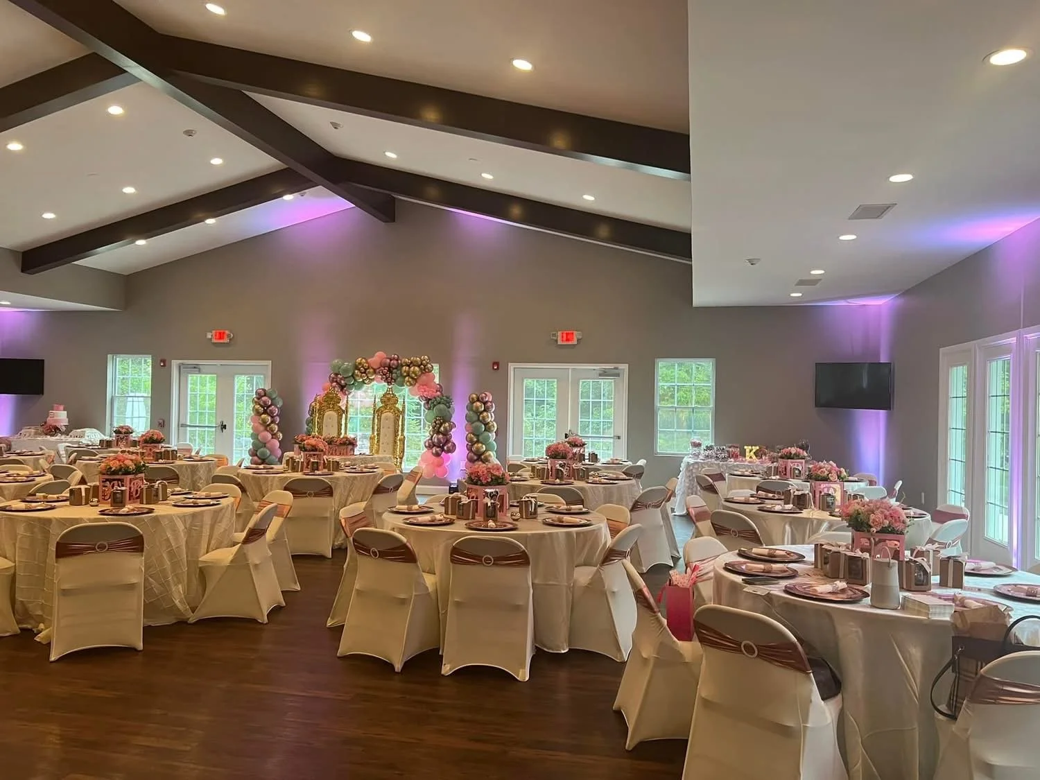 Decorated banquet hall with round tables, pink floral centerpieces, balloon arrangements, and gold accented chairs for a celebration event.
