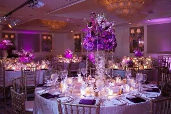 Elegant banquet room decorated with purple and pink floral centerpieces, candles, and purple lighting.