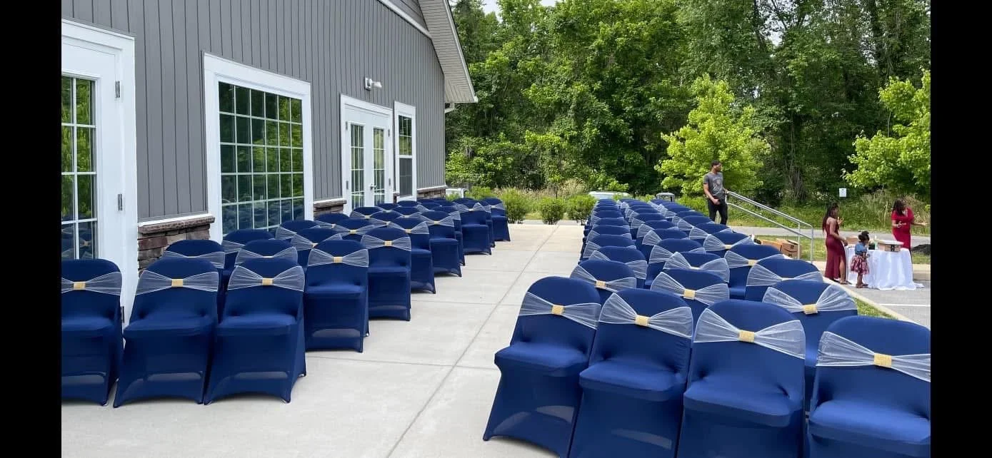 Rows of blue chairs with white decorative bows lined up outside a building with gray walls and large windows, preparing for an outdoor event.