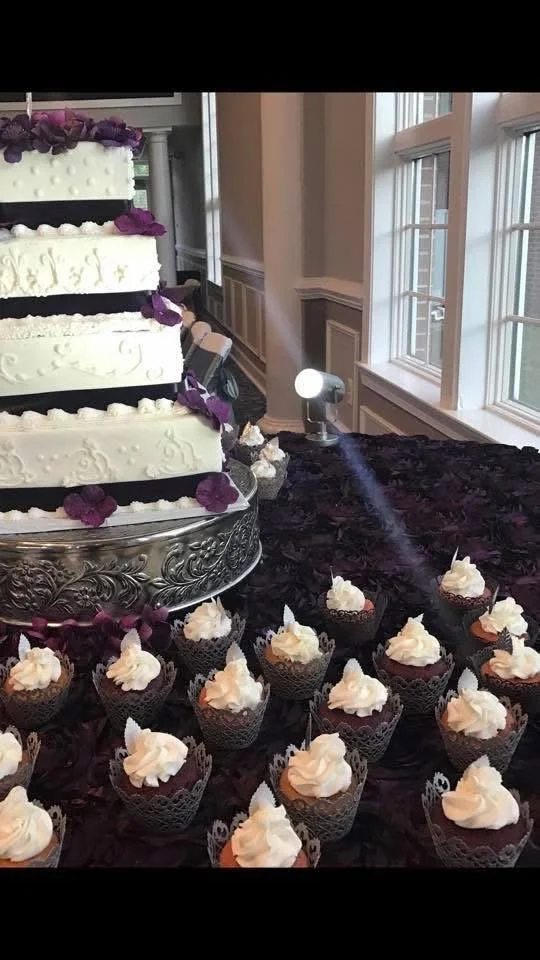 A wedding cake with white icing and purple flower decorations on a silver stand, with cupcakes topped with white frosting arranged in front of the cake on a dark tablecloth near windows.