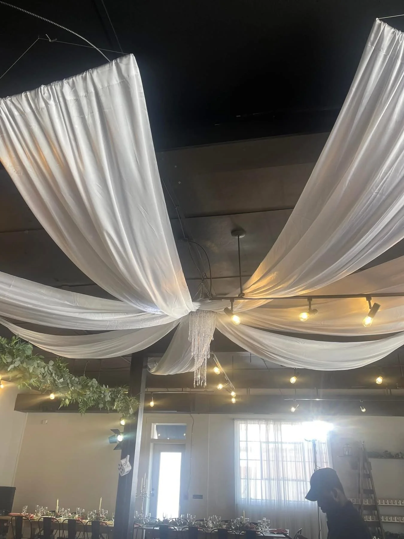 Interior of a decorated room with white draped fabric on the ceiling, string lights, a chandelier, and a dining area with tables set for an event.