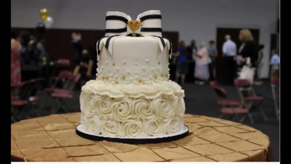 Two-tier wedding cake with white frosting, rosette decoration, a black and white striped bow with a bear topper, on a wooden table, wedding reception in the background.