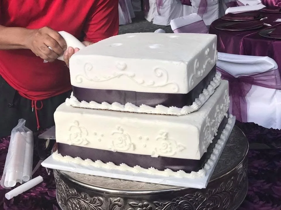Two-tiered wedding cake with white and purple decoration, ornate piping, sitting on a decorative stand at a celebration event.