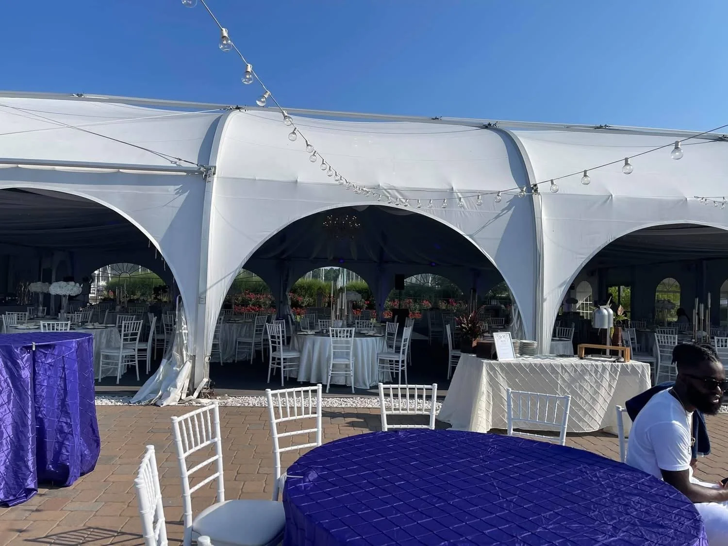 Outdoor event setup with white tents, white chairs, and tables with purple and white tablecloths under string lights on a sunny day.