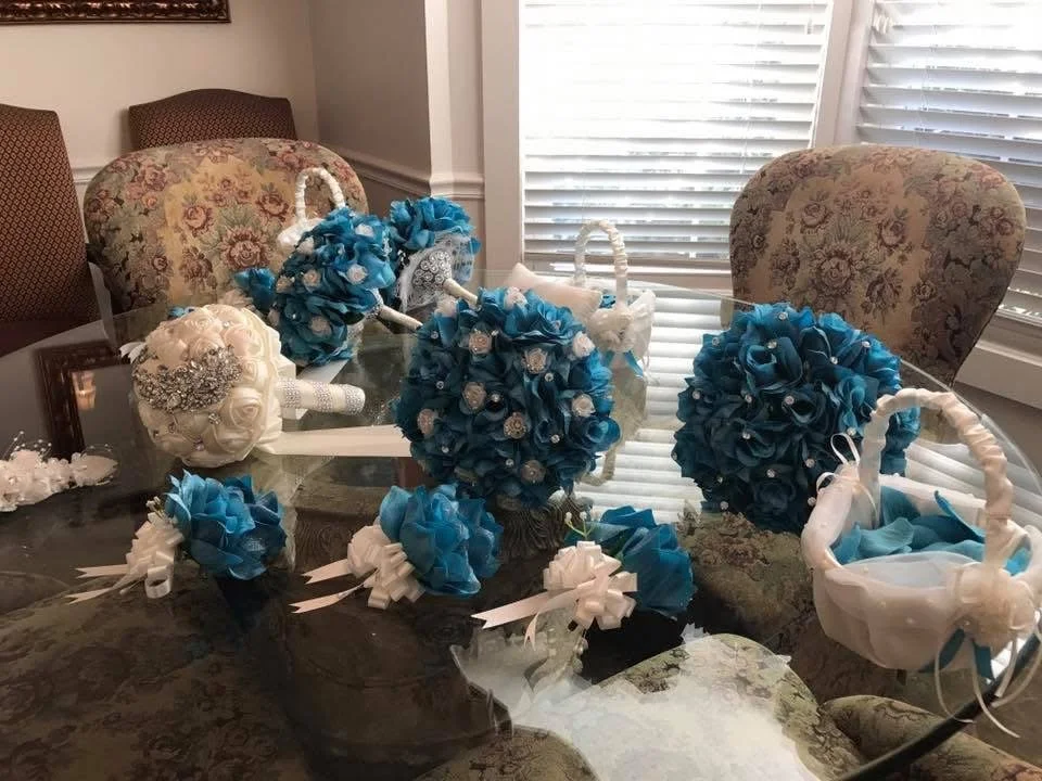 Bouquets and decorative accessories on a glass dining table, with chairs and window blinds in the background.