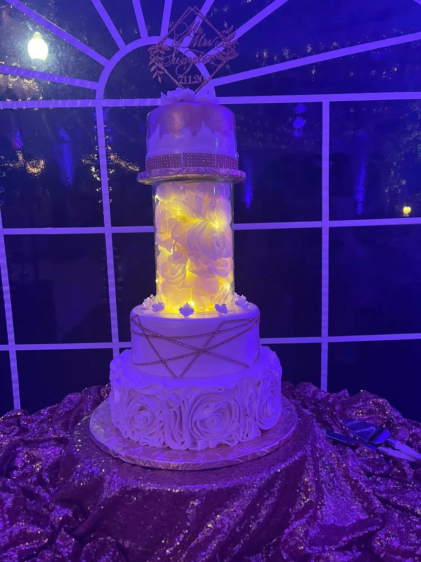 Four-tiered wedding cake with white icing and gold beads, topped with a cake topper, displayed on a purple sequin tablecloth with a backdrop of purple lighting.