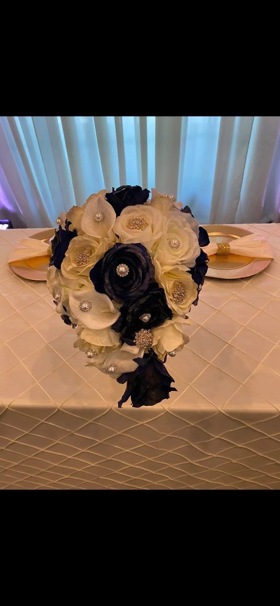 A bouquet of black and white flowers with decorative pearl and rhinestone embellishments, placed on a table with a white quilted tablecloth. In the background, there are two plates with napkins and a gold-colored trim, and sheer curtains let in soft 