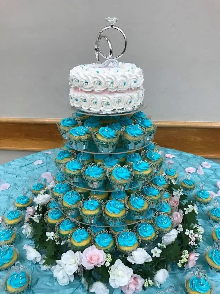 A wedding cake topped with silver rings and surrounded by blue cupcakes with blue swirl frosting, all arranged on a tiered stand and decorated with white and pink flowers.