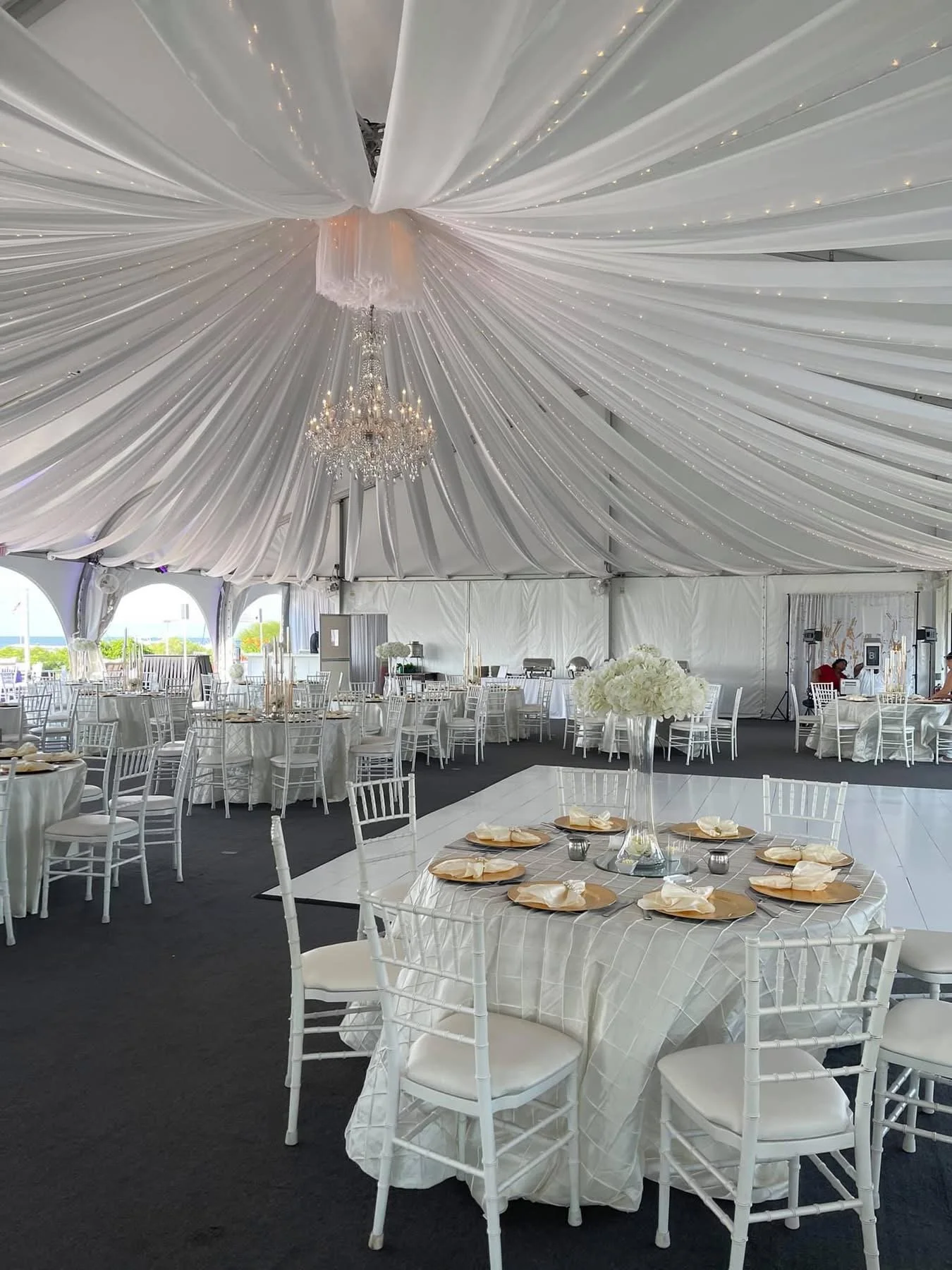 Interior of a decorated banquet tent with white drapes, a chandelier, and set tables with white chairs and floral centerpieces.