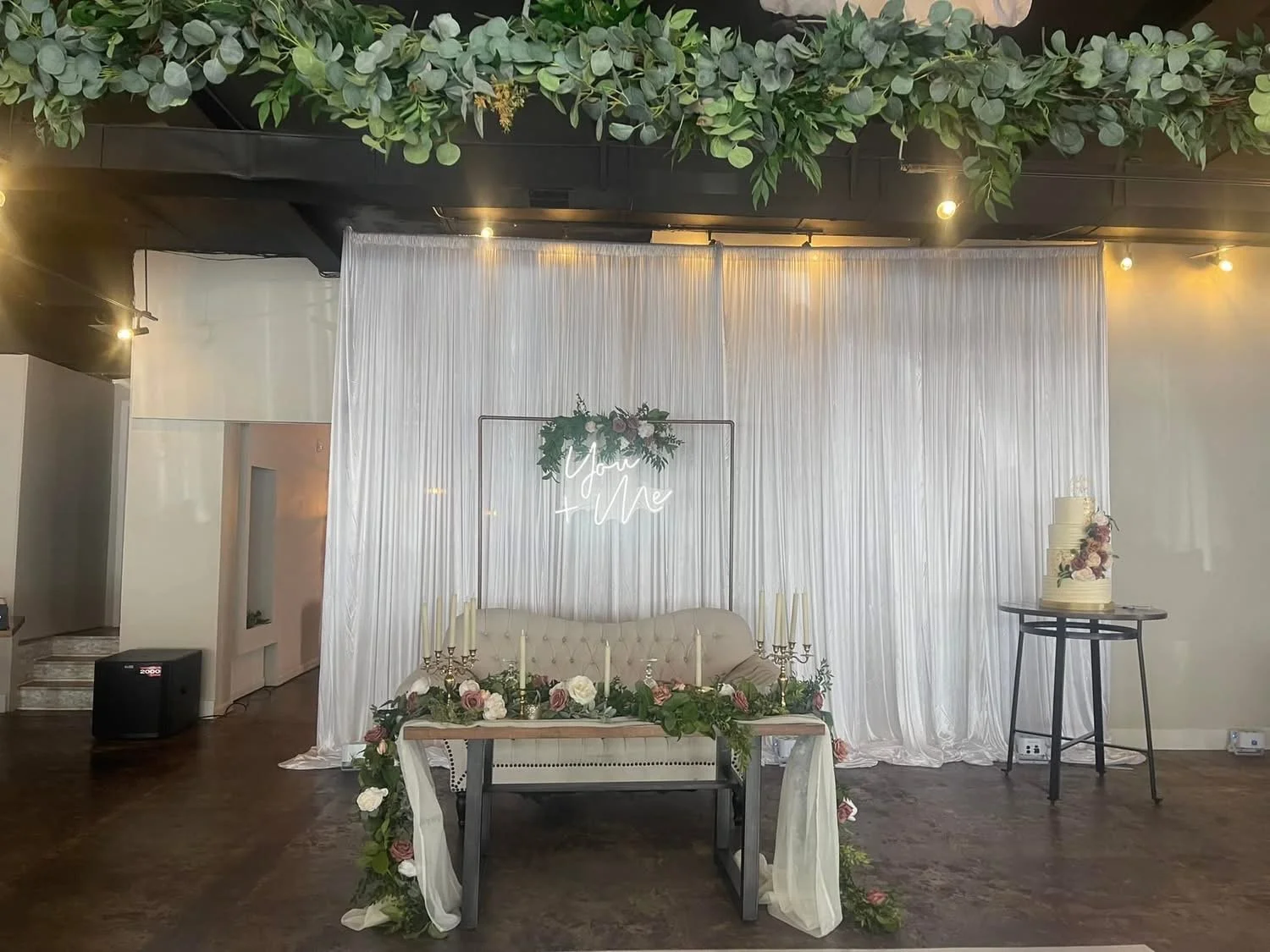Wedding celebration setup with a white backdrop, floral arrangements, a sign that says "You & Me," a table with candlesticks and flowers, a sofa, and a wedding cake on a stand.