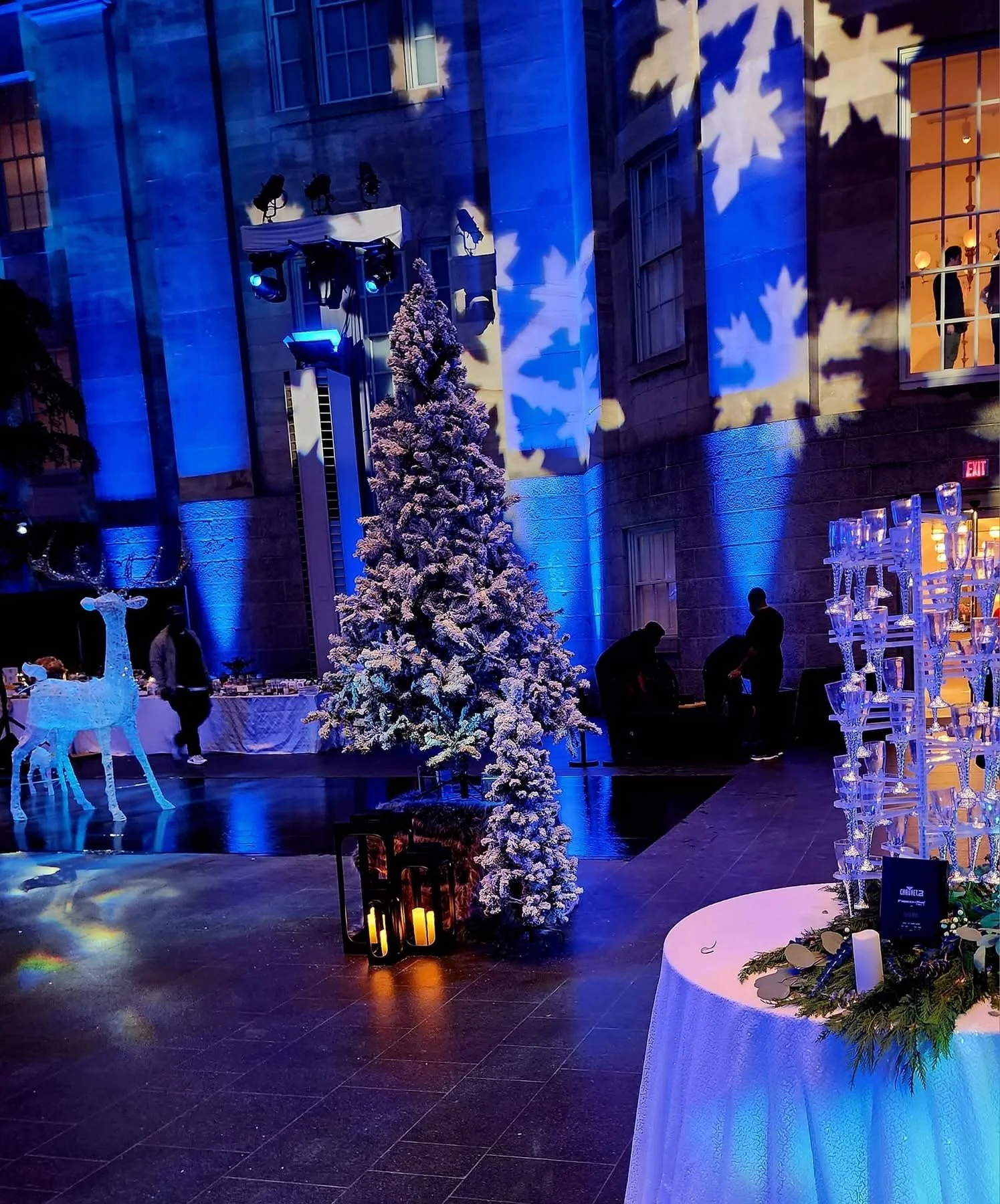 A decorated indoor space featuring a Christmas tree with snow-like decorations, illuminated candles in lanterns on the floor, and a display of clear glassware arranged on a round table. There are people in the background, and snowflake and winter-the