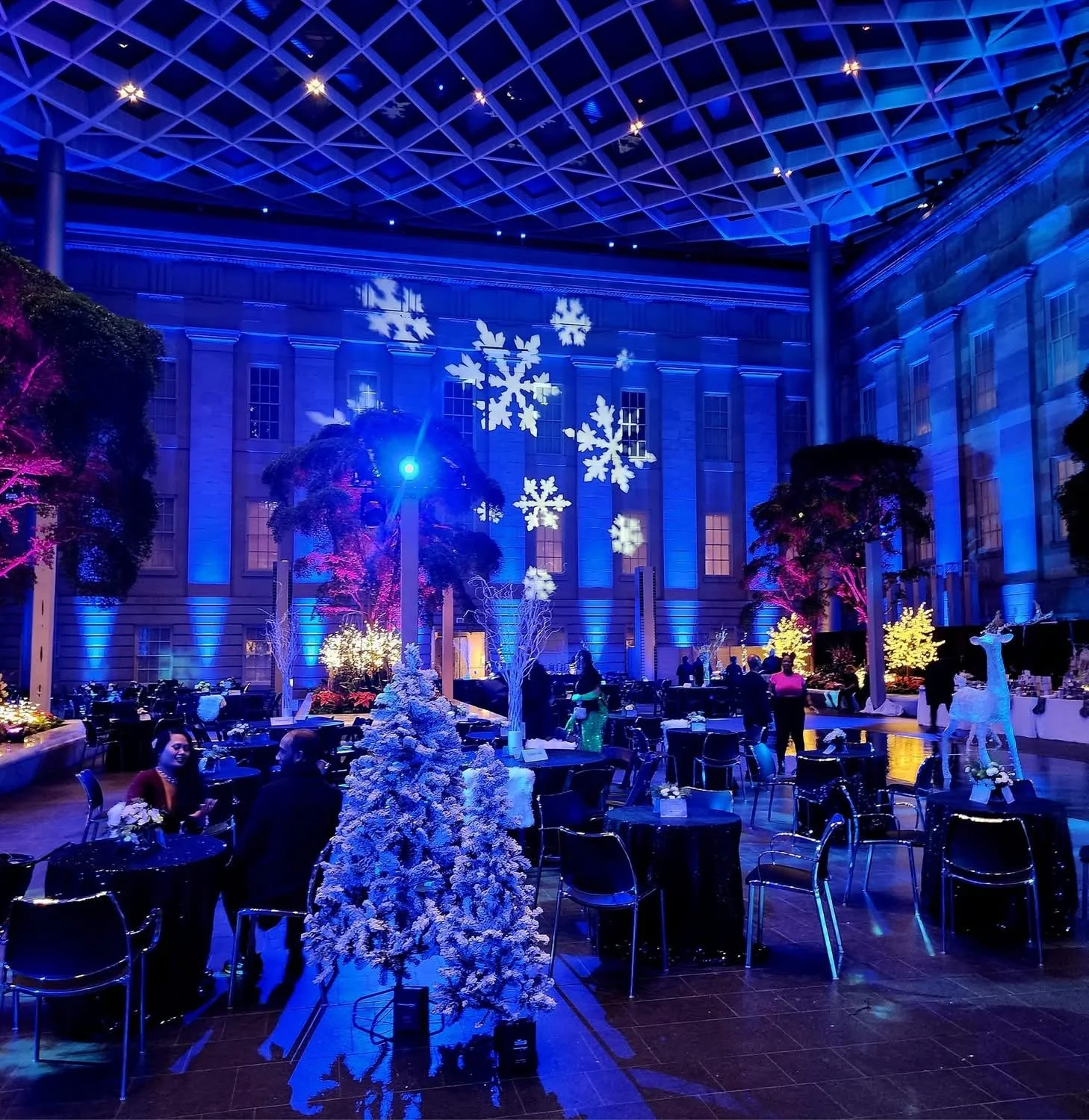 Indoor event space decorated with snowflakes projected onto the walls, illuminated trees, and festive holiday decorations, with people socializing at round tables, including a white reindeer figurine.