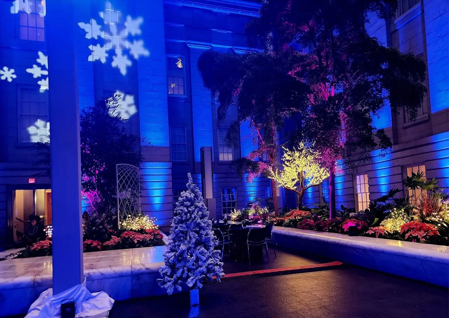 Indoor courtyard decorated with colorful holiday lights and ornaments, including snowflake projections on the wall, illuminated trees, and festive plants.
