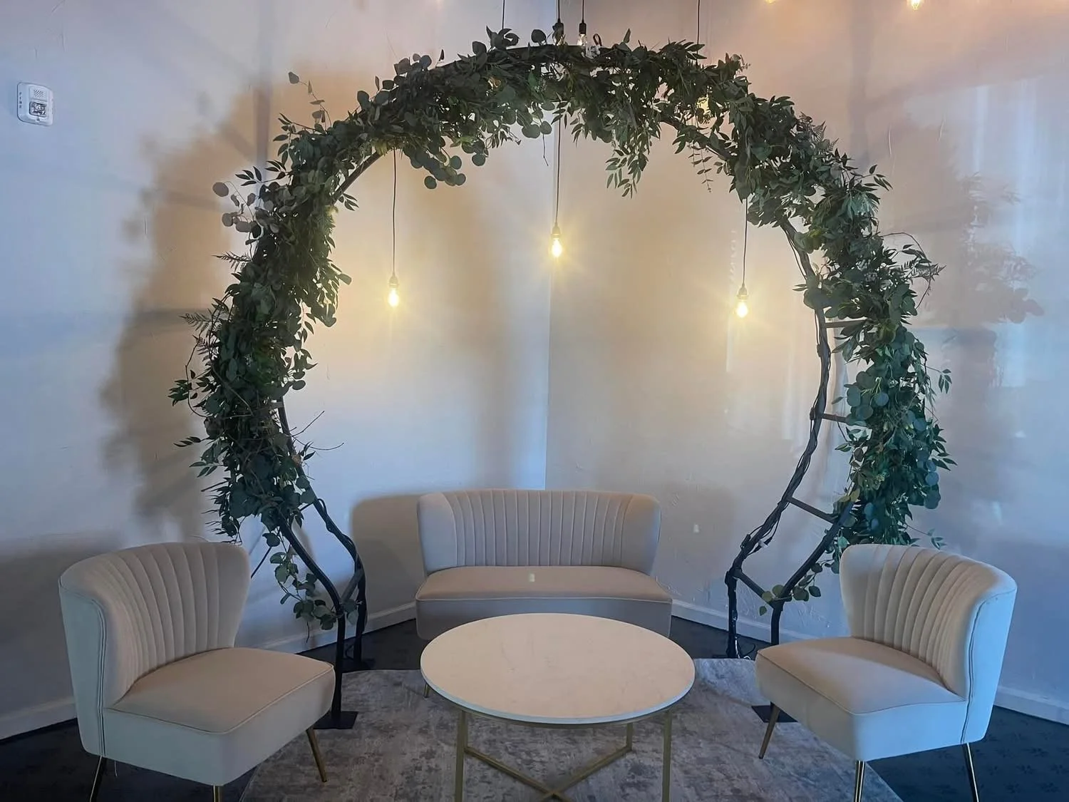 A cozy seating area with three white, vintage-style chairs and a white round table, set against a plain wall. A decorative arch made of greenery and twigs stands behind the chairs, adorned with three hanging Edison-style light bulbs producing warm li