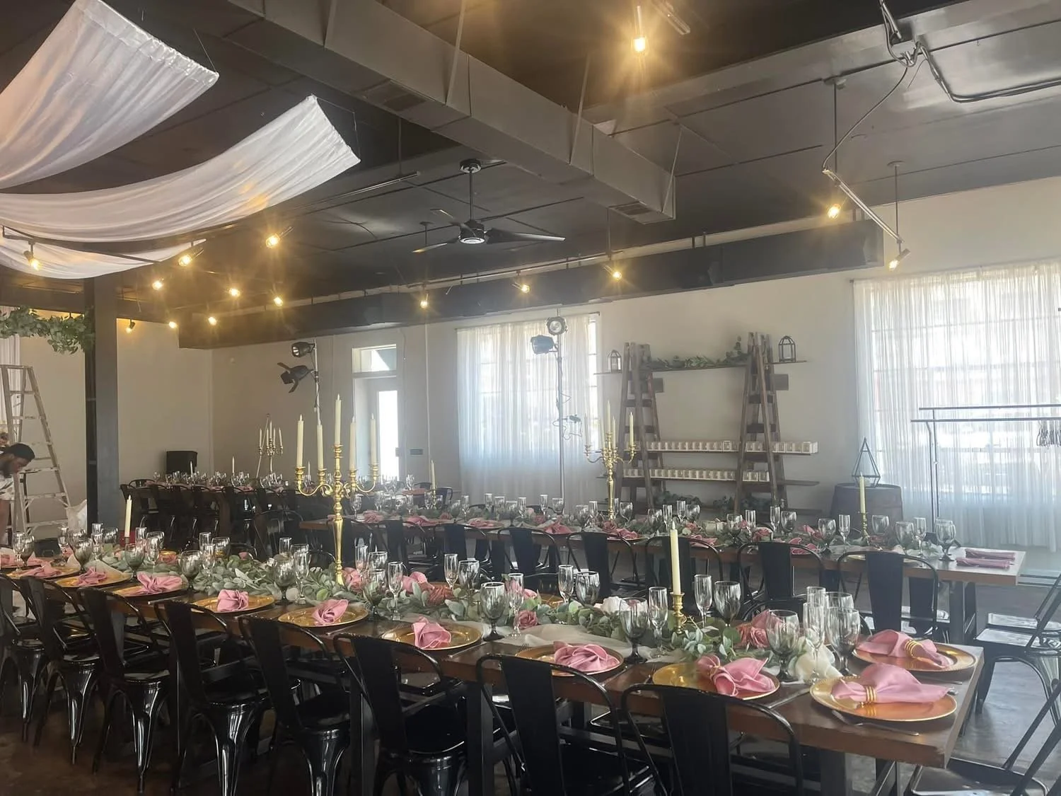 A large dining table set for a formal event with pink napkins, gold chargers, glassware, and floral centerpieces, inside a decorated room with curtains, chandeliers, and stage lighting.