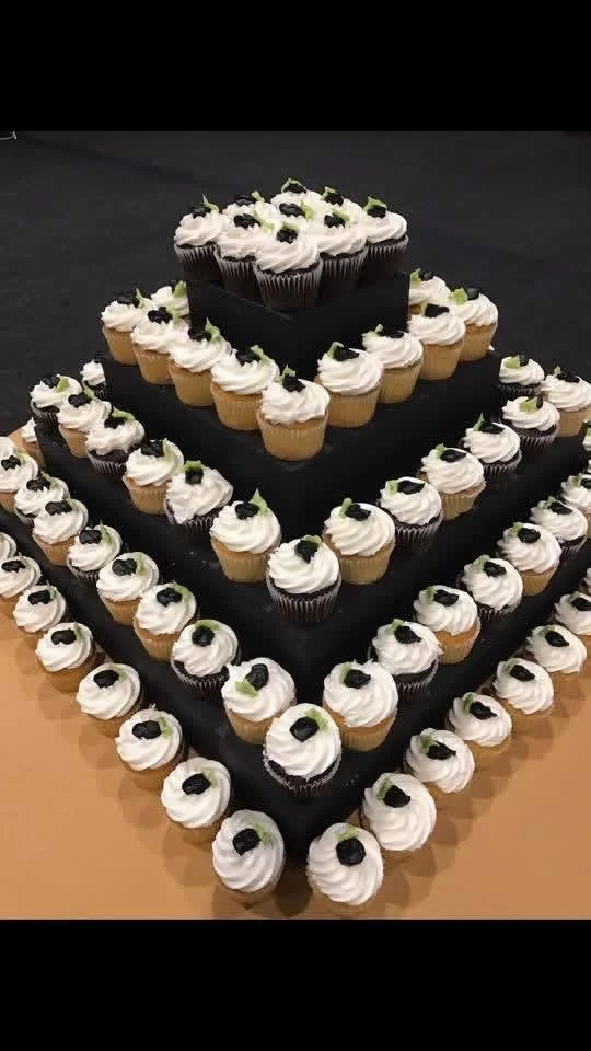 A large, multi-layered cupcake cake with white frosting, topped with small black and green decorations, arranged in a tiered square shape on a black surface.