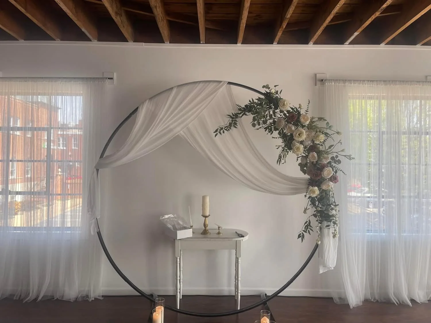 Circular metal arch decorated with white and blush pink flowers and greenery, draped with white fabric, set against white walls and sheer curtains, with candles on the floor and a small white table underneath.
