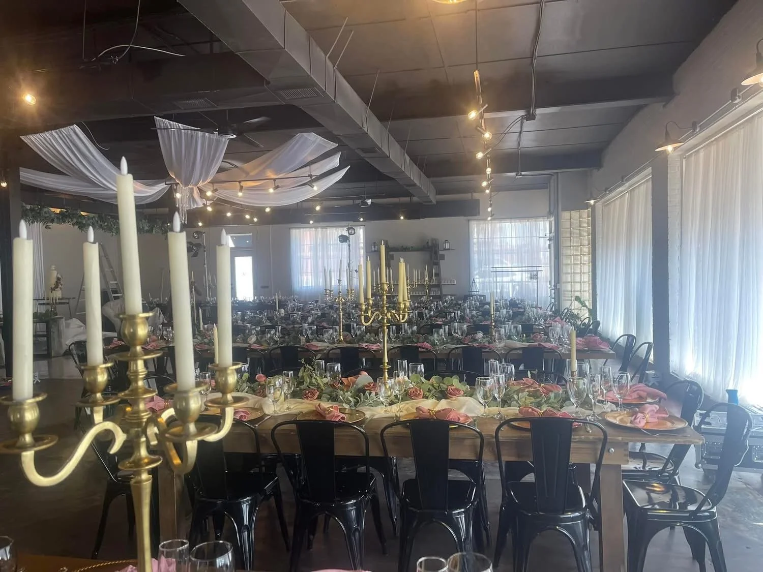 Wedding reception setup with long tables, pink napkins, floral centerpieces, glassware, and gold candelabras in a decorated banquet hall.