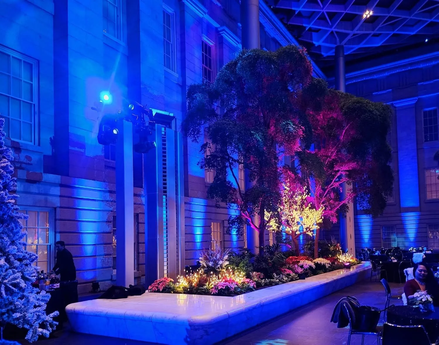 Indoor event space with colorful lighting illuminating a large tree and surrounding plants, tables set for dining, and a person preparing drinks.