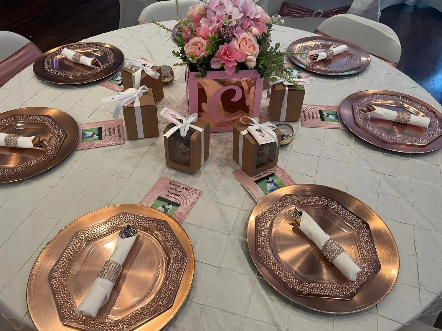 A round table decorated with pink and gold themed centerpieces, pink flowers in a pink box, gold gift boxes with white ribbons, pink and white chance cards, gold plates with white napkins and silverware, and small decorative items for a celebration.