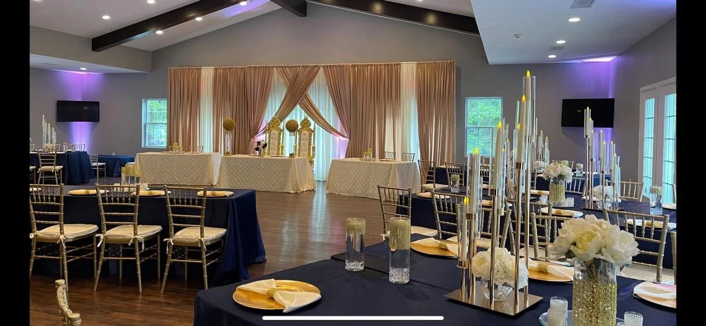 Elegant banquet hall decorated for a wedding reception with tables adorned with floral centerpieces, tall candles, and gold accents. Draped curtains and purple lighting create a sophisticated ambiance.