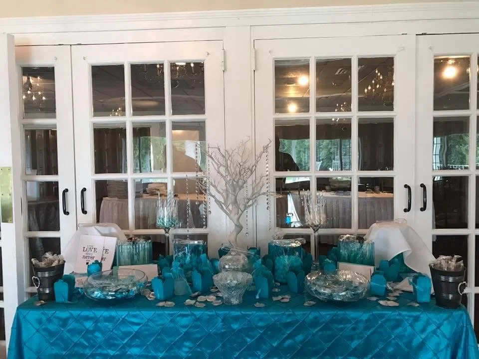 Decorative table setup with blue themed items, including a blue quilted tablecloth, glass vases, and teal gift bags, in front of white framed glass doors.