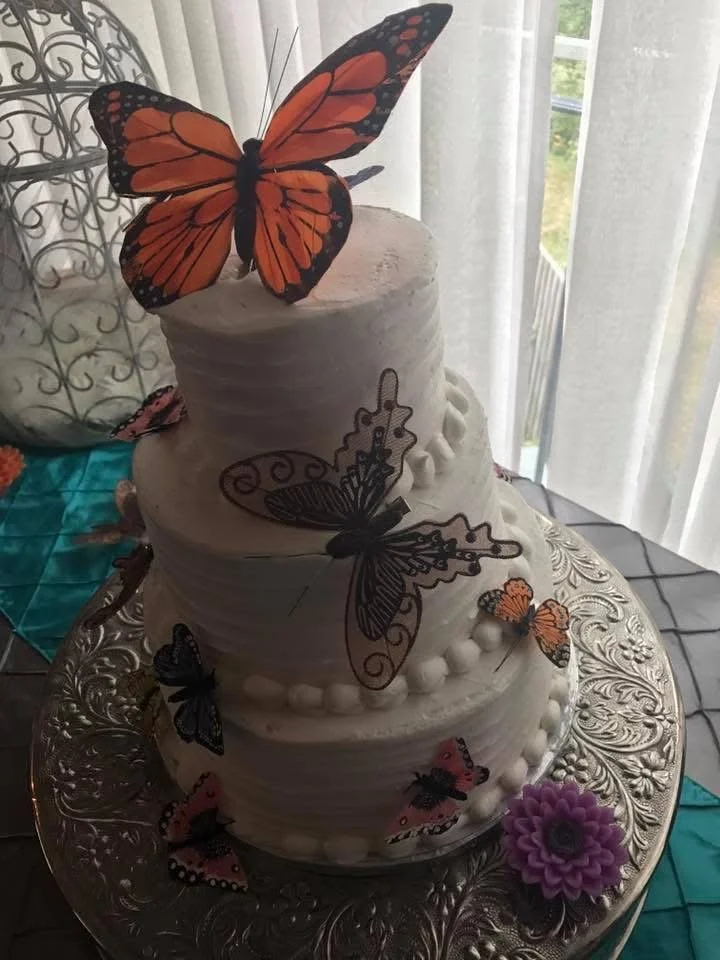 Three-tiered white birthday cake decorated with edible butterflies and pearl-like accents, displayed on an ornate silver stand near a window with white curtains.