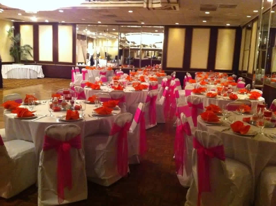 Event space decorated with round tables covered in white tablecloths, pink chair sashes, and orange napkins, set for a celebration or banquet.