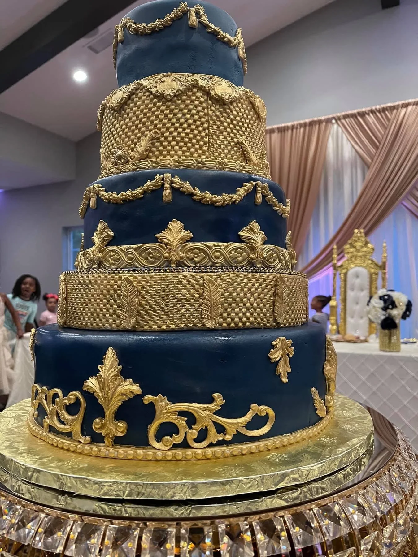 A multi-tiered cake with blue and gold decorations, featuring gold ornate patterns, garlands, and fondant embellishments, displayed on a gold and crystal stand at a celebratory event.