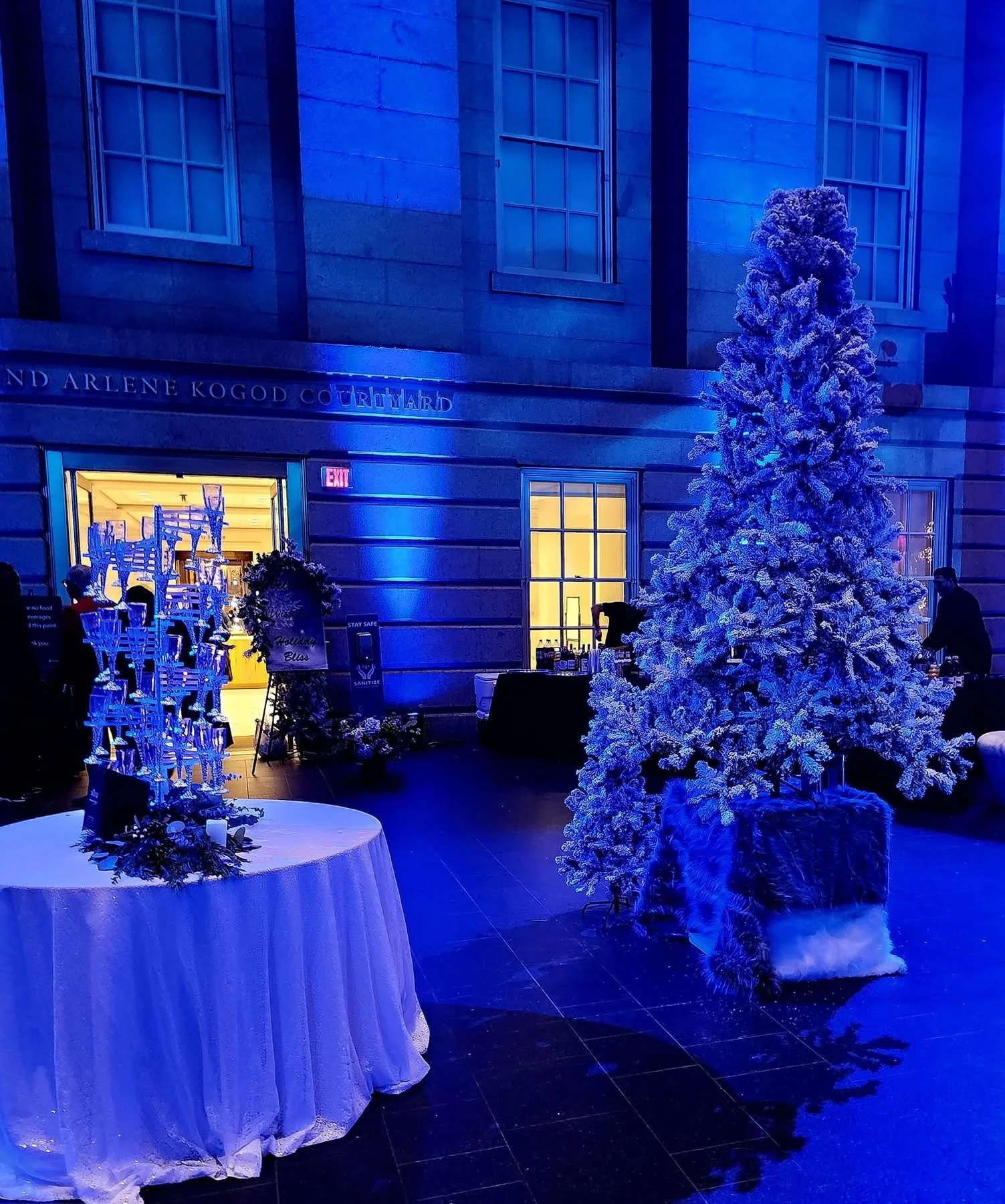 Decorated Christmas tree with snow-like white flocking, illuminated by blue lighting, inside a building with string lights, a table with a white cloth, and a photo display stand