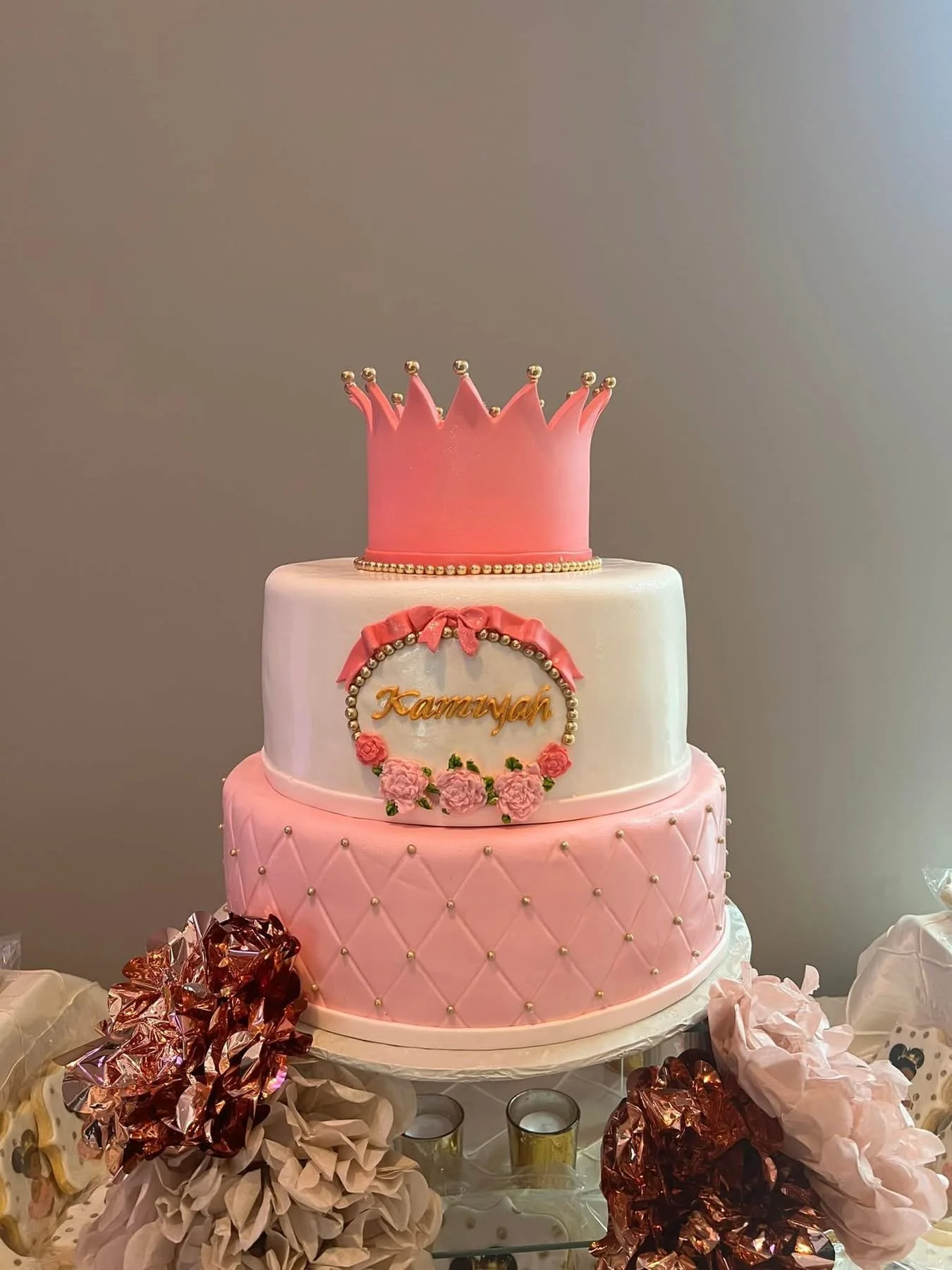 Three-tiered pink and white birthday cake decorated with a crown on top, pink flowers, and the name 'Kamiyah' on the middle tier, surrounded by pink, gold, and brown decorations.