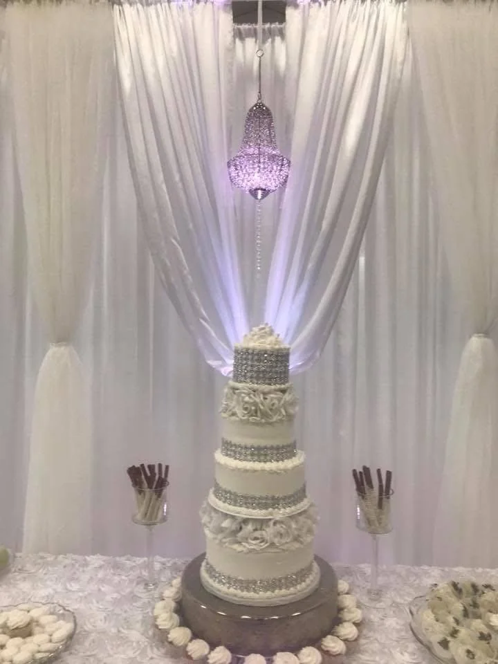 A multi-tiered white wedding cake decorated with silver embellishments and flowers, placed on a table with white floral tablecloth. Behind the cake, there are white curtains with purple lighting, and a pink chandelier hanging from the ceiling. There 