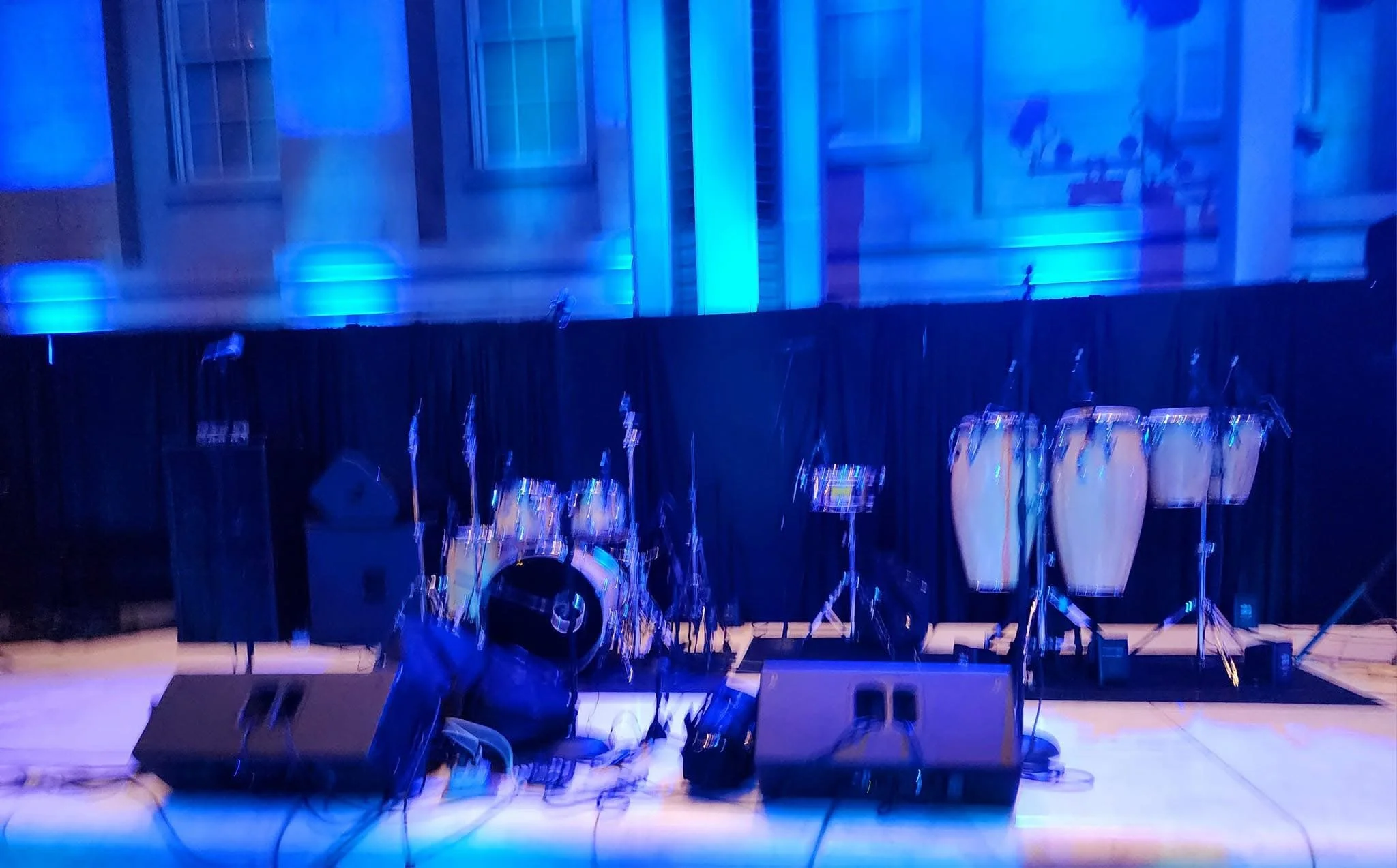 An empty stage with musical instruments including conga drums, a drum set, microphones, and speakers, illuminated with blue lighting and set against a black curtain backdrop.