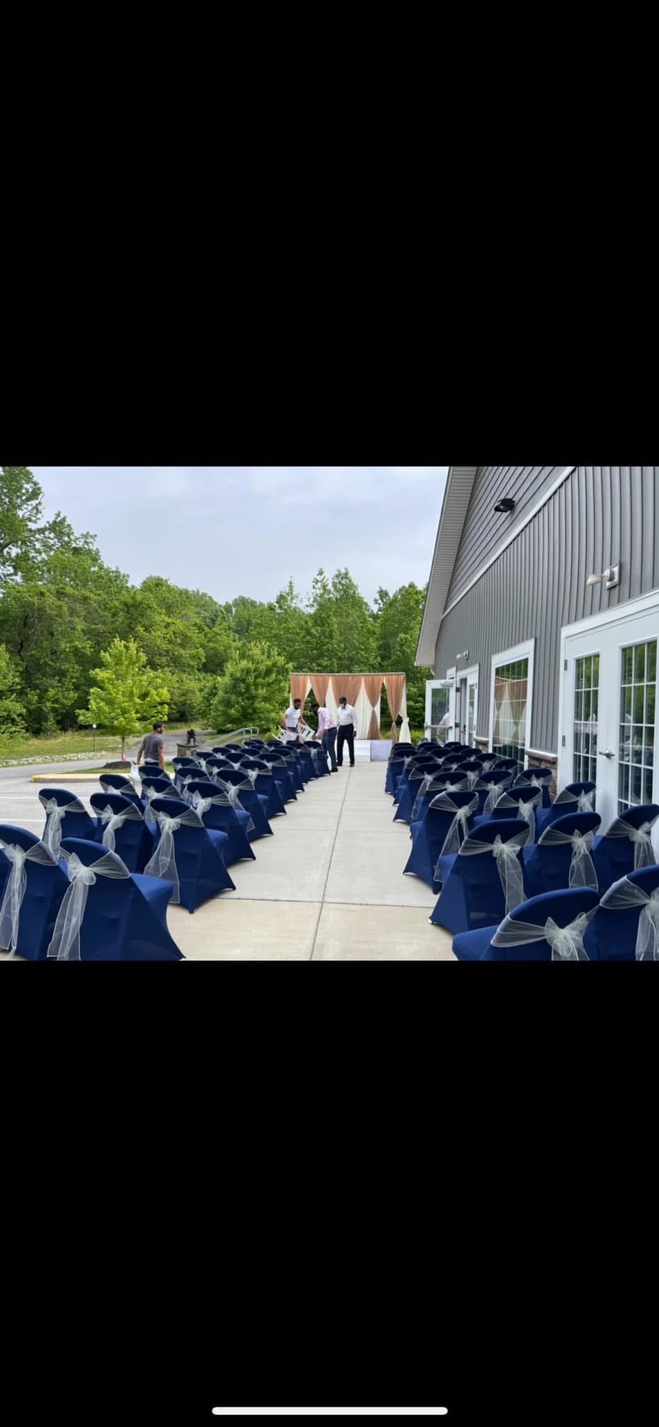 Outdoor event setup with rows of decorated navy blue chairs with white bows, facing a stage at a venue near a forest, overcast sky.