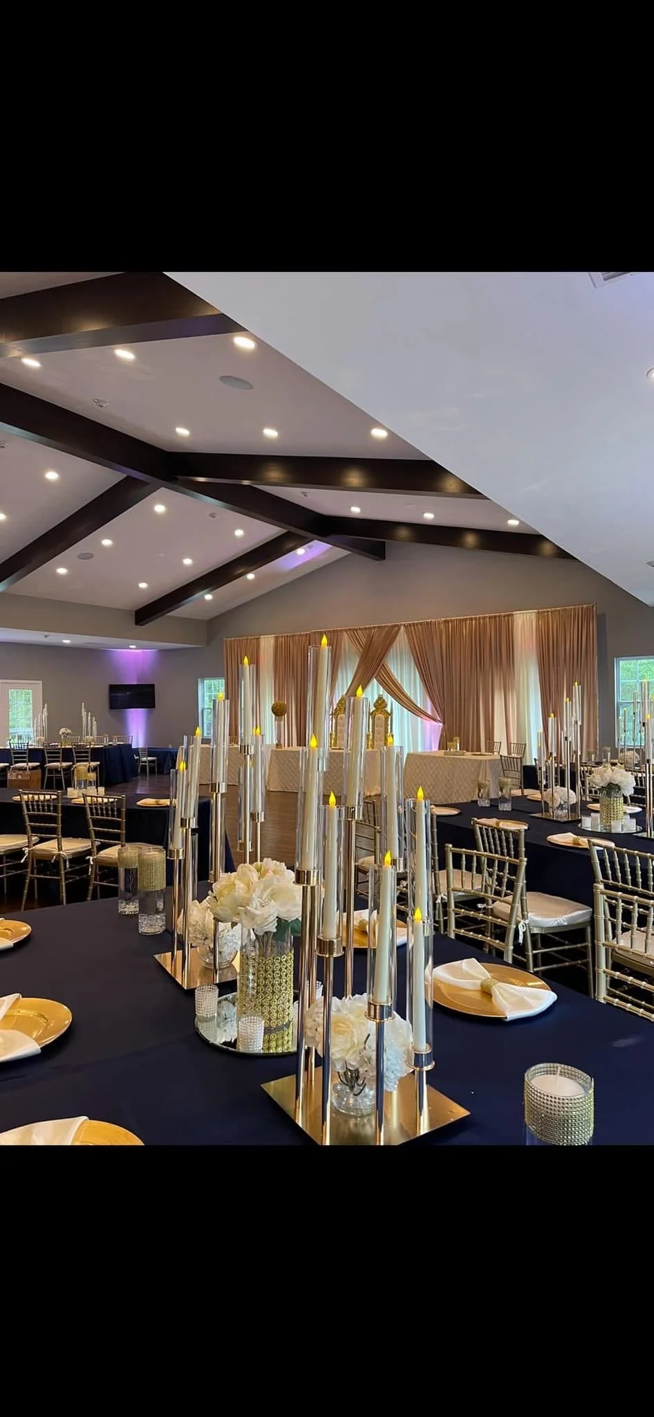 Elegant event reception hall decorated with long tables, gold accents, tall candles, white floral centerpieces, and draped curtains in the background.