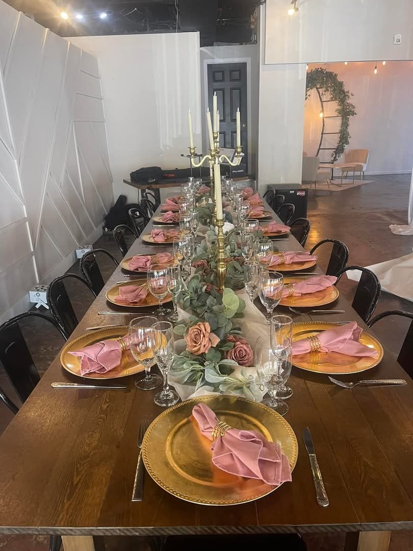 A long wooden dining table set for a formal event with gold chargers, pink napkins, glassware, and a gold candelabra centerpiece with pink roses and greenery. The background shows a modern interior with white walls and a seating area.