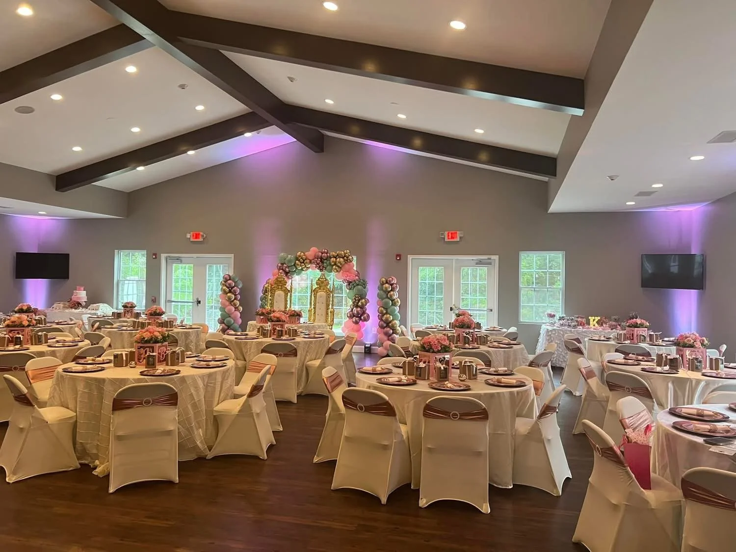 Decorated banquet hall with pink and gold accents, round tables with pink floral centerpieces, balloon arch and backdrop in pink, purple, and gold, set for a celebration or party.