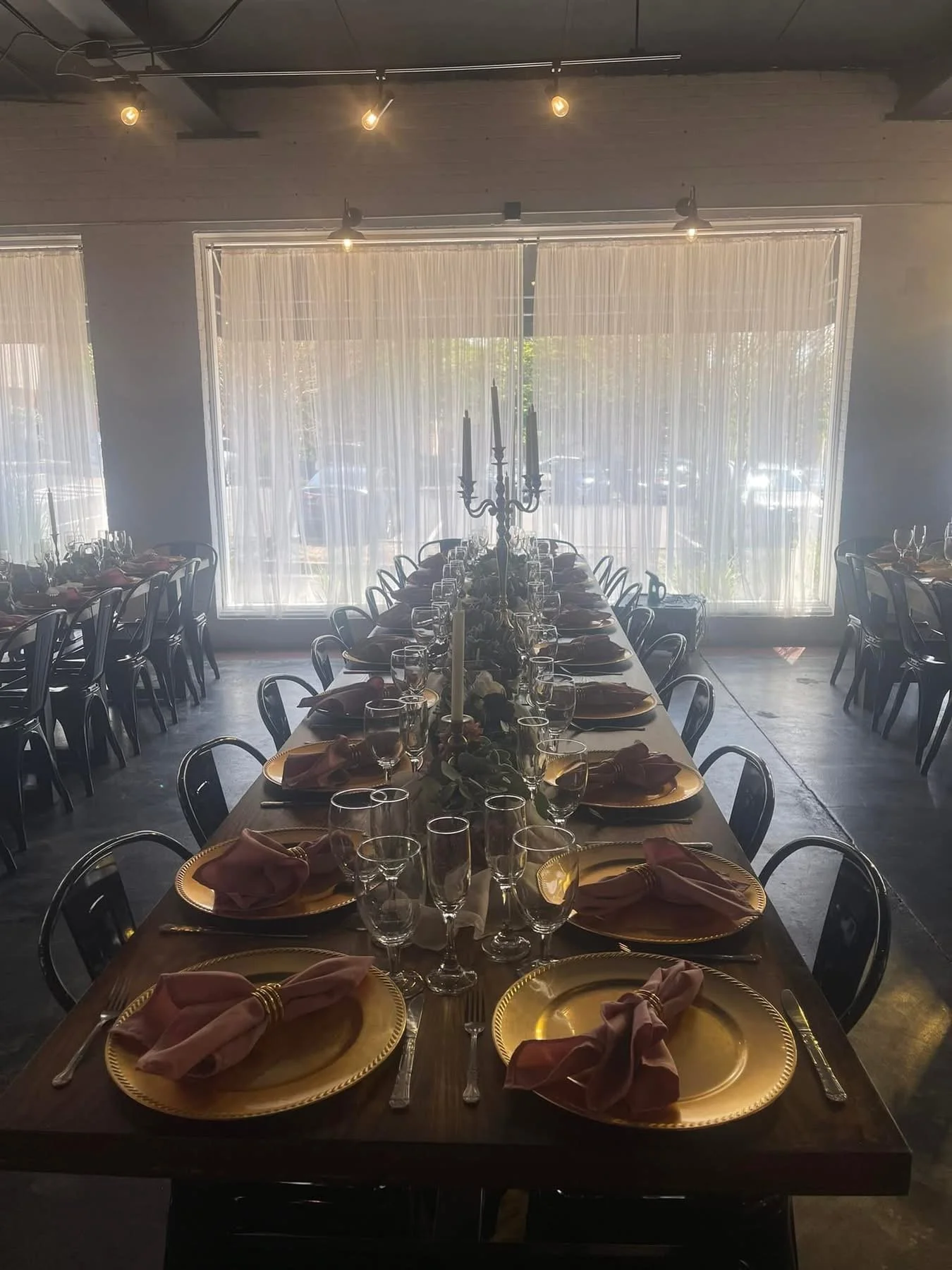 A long rectangular dining table set for a formal event with gold chargers, pink napkins, wine glasses, and silverware, inside a bright room with large windows and sheer curtains.