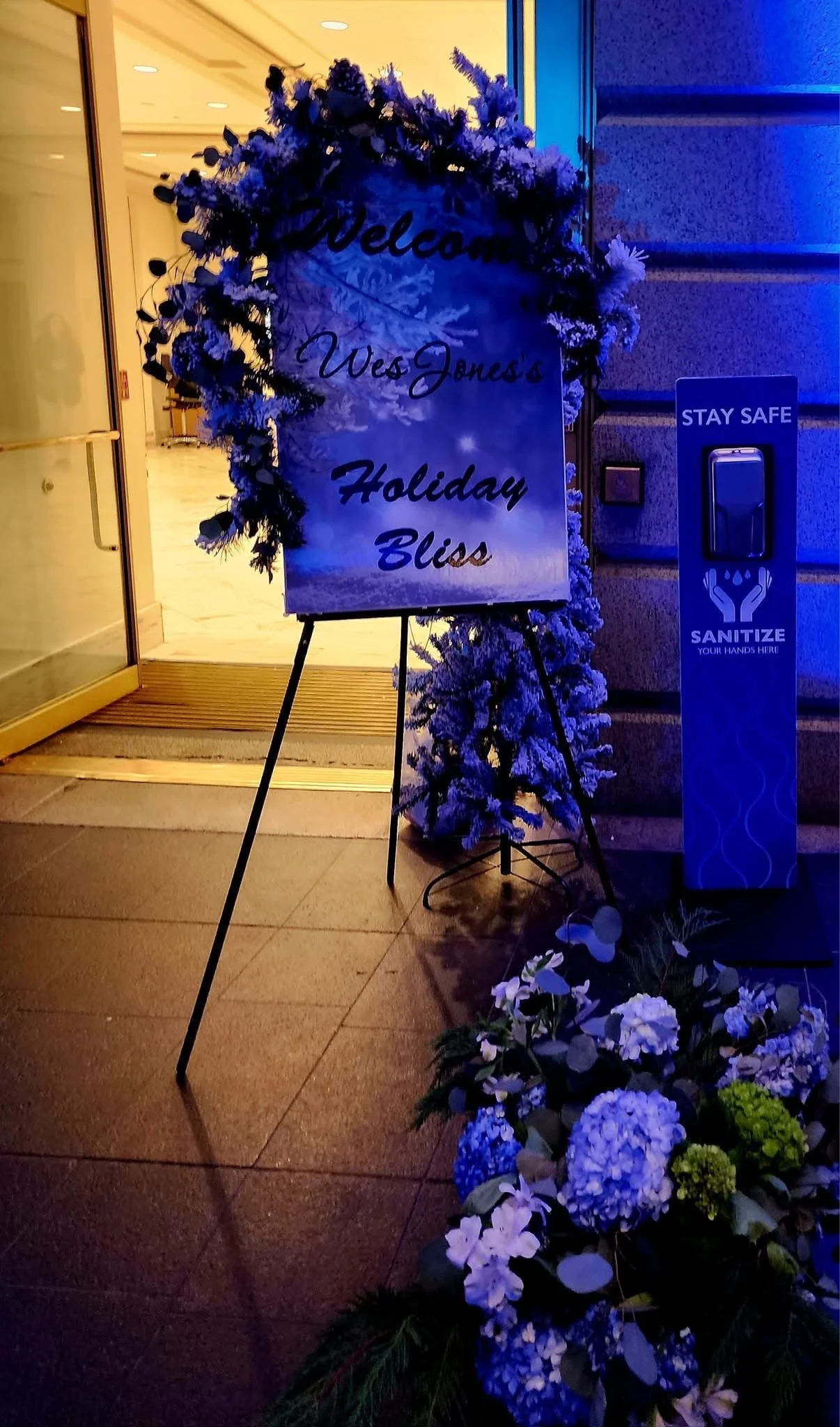 A welcome sign decorated with a purple floral wreath reads 'Welcome Wes Jones's Holiday Bliss'. The sign is near an entrance with an open door. To the right, there is a blue hand sanitizer station with the words 'STAY SAFE' and 'SANITIZE YOUR HANDS H