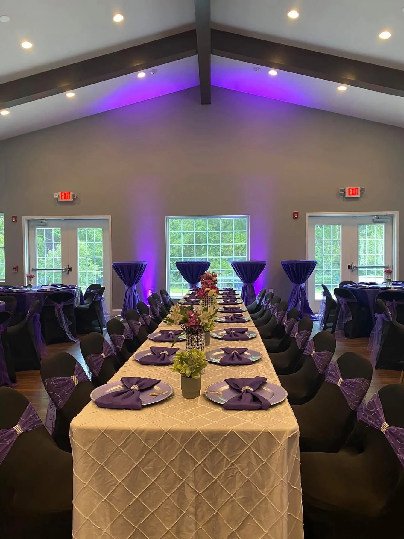Wedding reception setup with a long head table, black chairs with purple bows, and round tables with similar decorations, purple uplighting, and floral centerpieces.