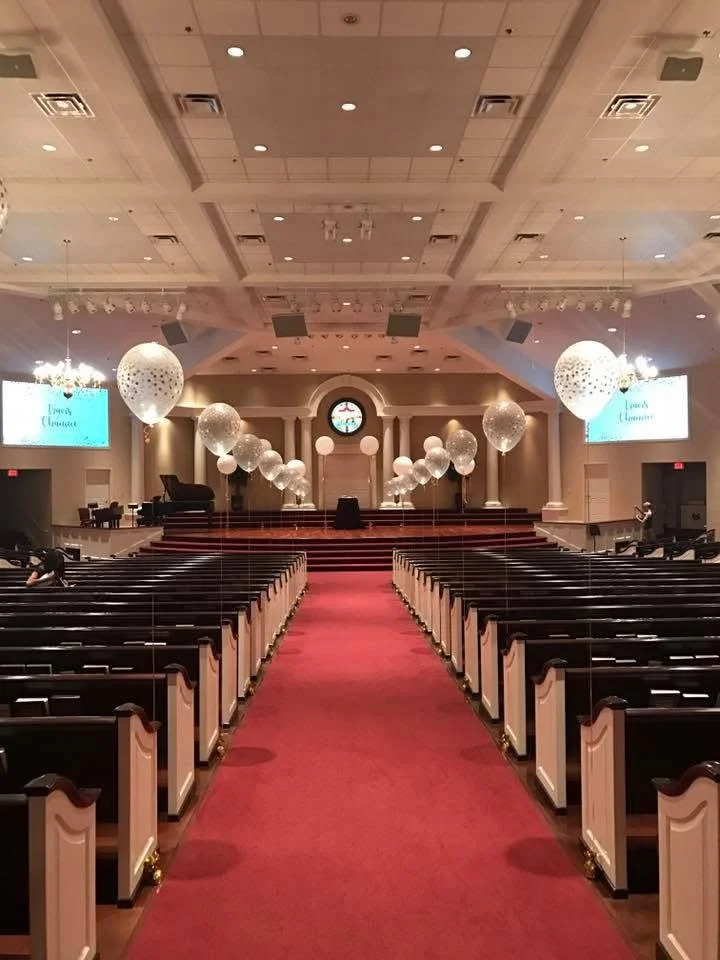Empty church or wedding hall decorated with large white balloons and a red carpet aisle leading to a stage with a clock and screens displaying 'Lindsay & Lance'.