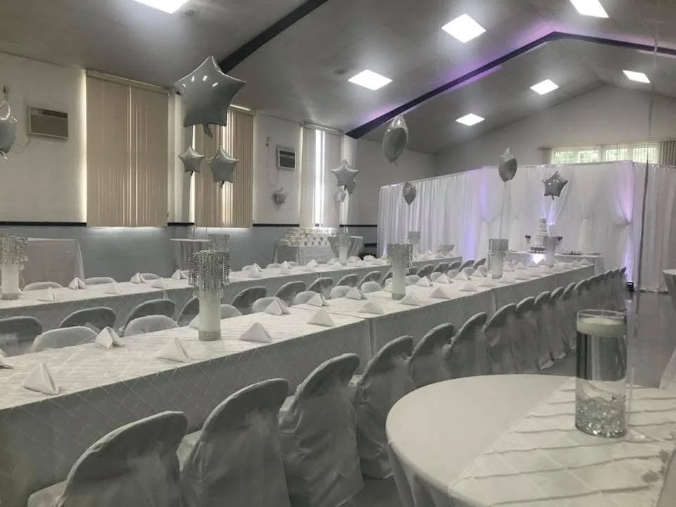 A banquet hall decorated for a celebration with white tablecloths, white chairs, and star-shaped balloons, featuring tall centerpieces with candles and crystal decorations.