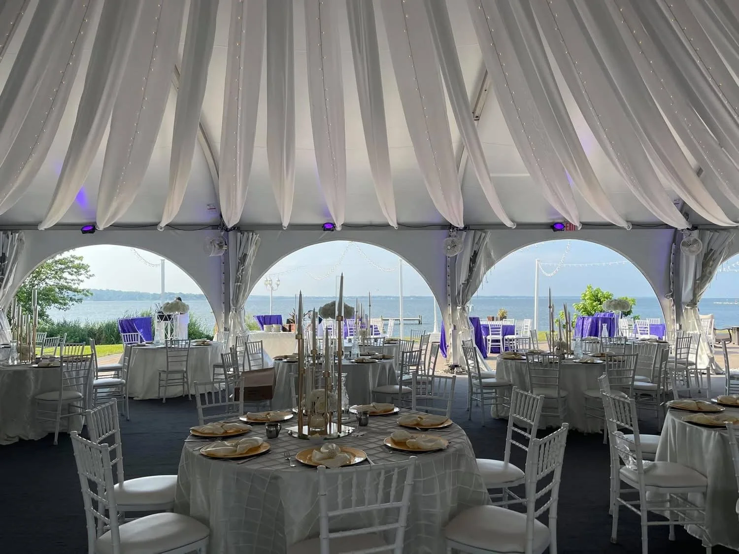 Elegant outdoor event setup with round tables covered in white tablecloths, decorated with folded napkins and centerpieces, under a tent with draped white fabric, overlooking a body of water with sailboats and greenery in the background.