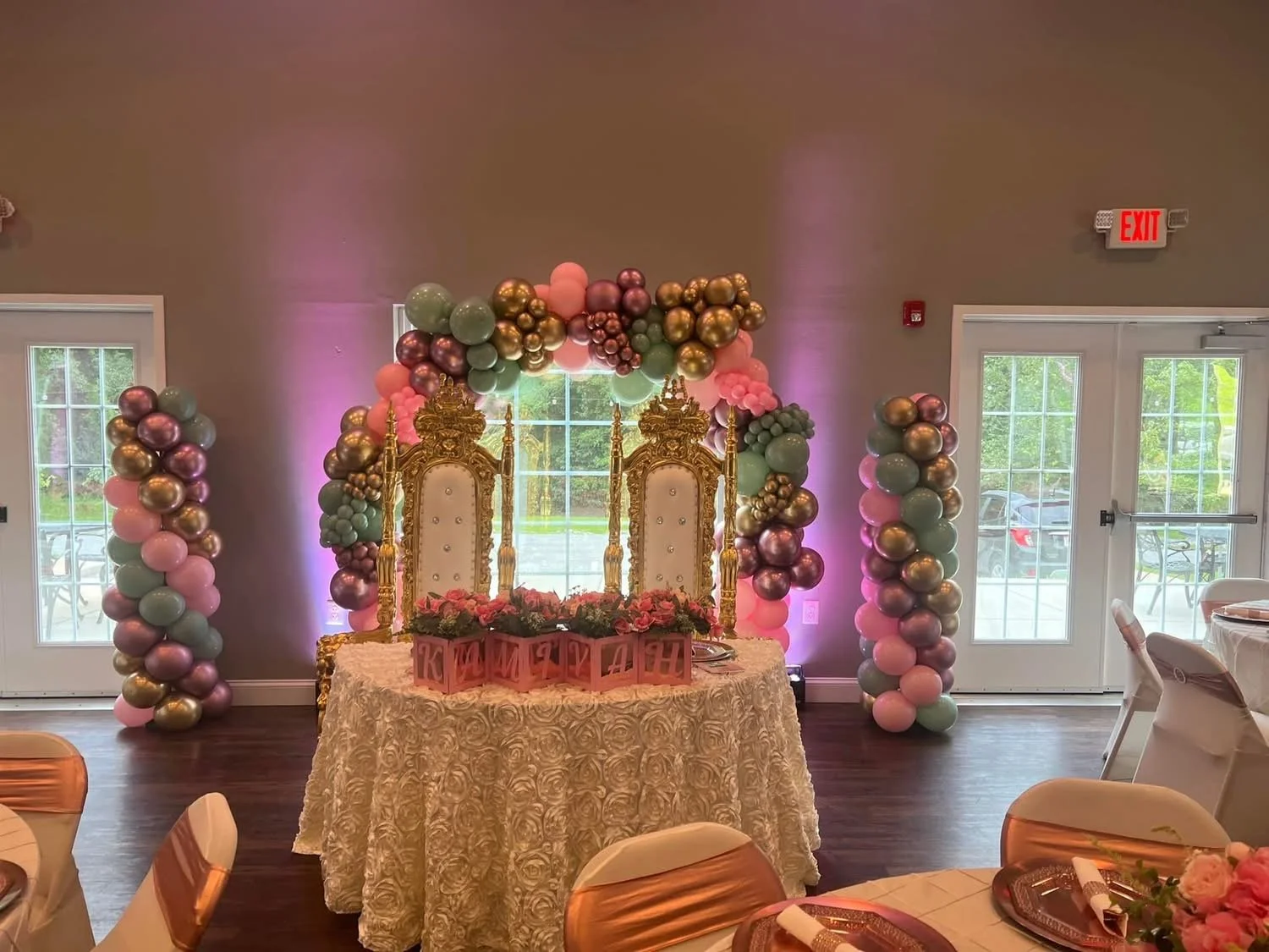 Elegant event setup with a decorated backdrop, golden thrones, and colorful balloon arch with pink, green, and metallic balloons, table with floral arrangements, and chairs with white covers and gold sashes.
