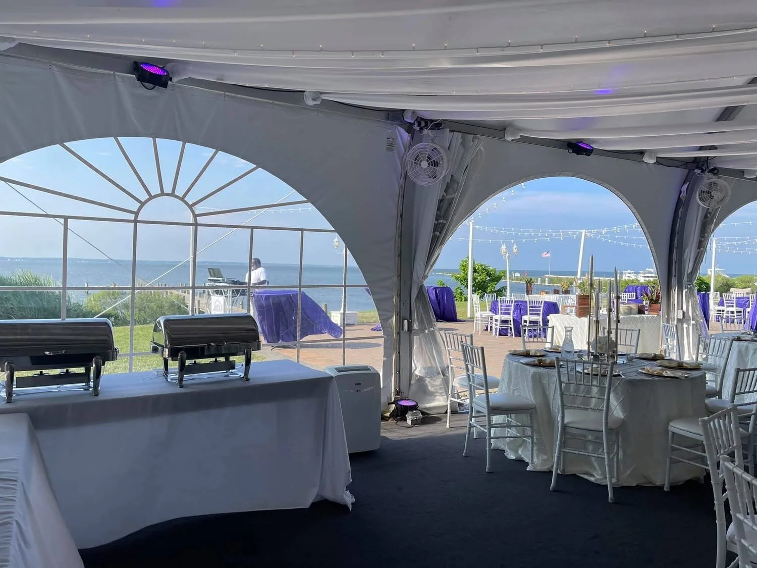Wedding reception tent overlooking a waterfront with decorated tables and chairs, buffet table, and a DJ or entertainer outside.